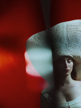 Woman in a large straw hat poses in front of a vibrant red backdrop, illuminated by soft, diffused lighting creating a surreal atmosphere.