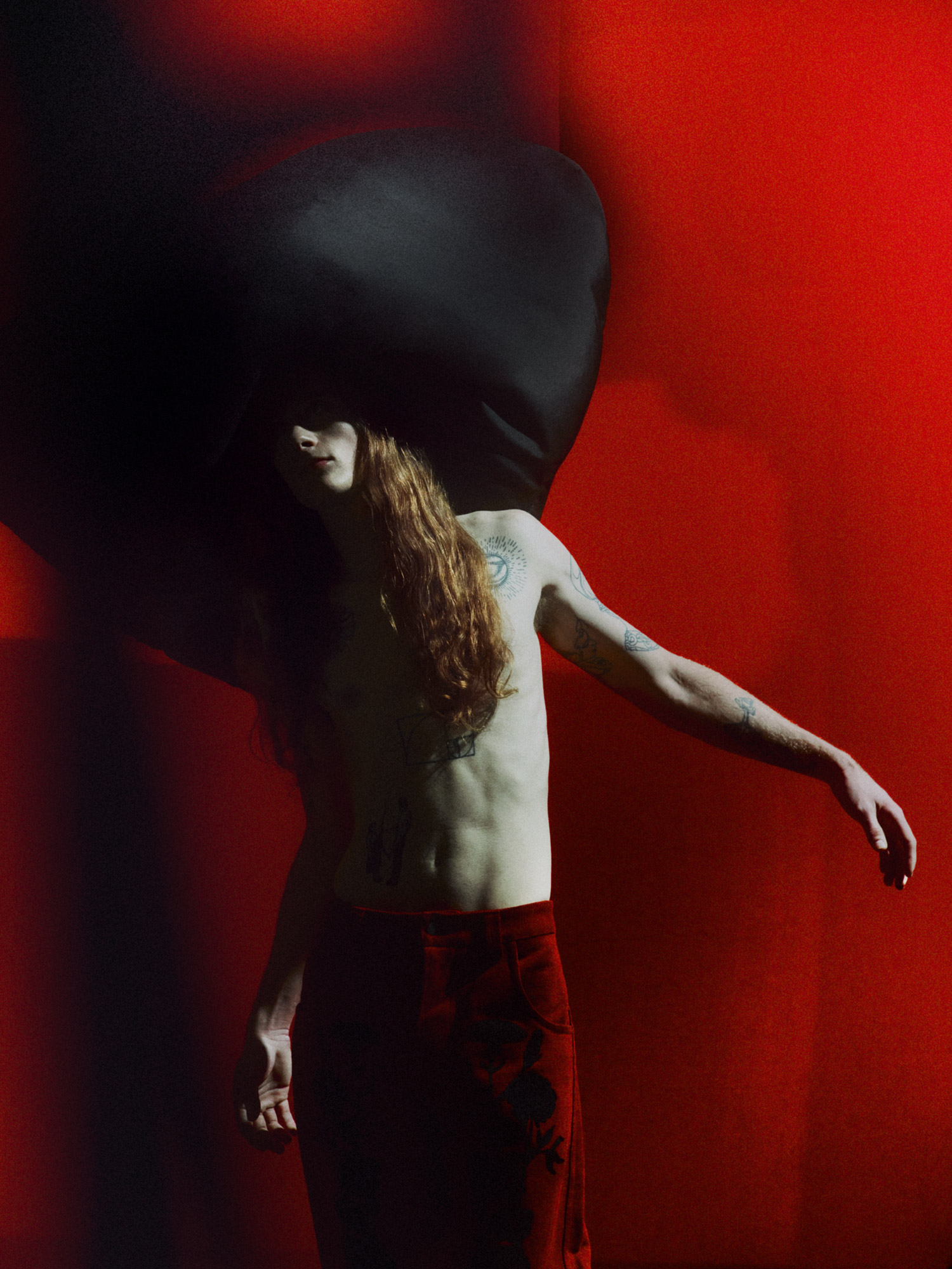 Adult male model with long red hair poses shirtless against a red wall with a black garment draped around him in a dramatic studio shot.