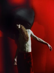 Shirtless model with long reddish hair wearing dark red trousers against a red background with a large black sculptural headpiece