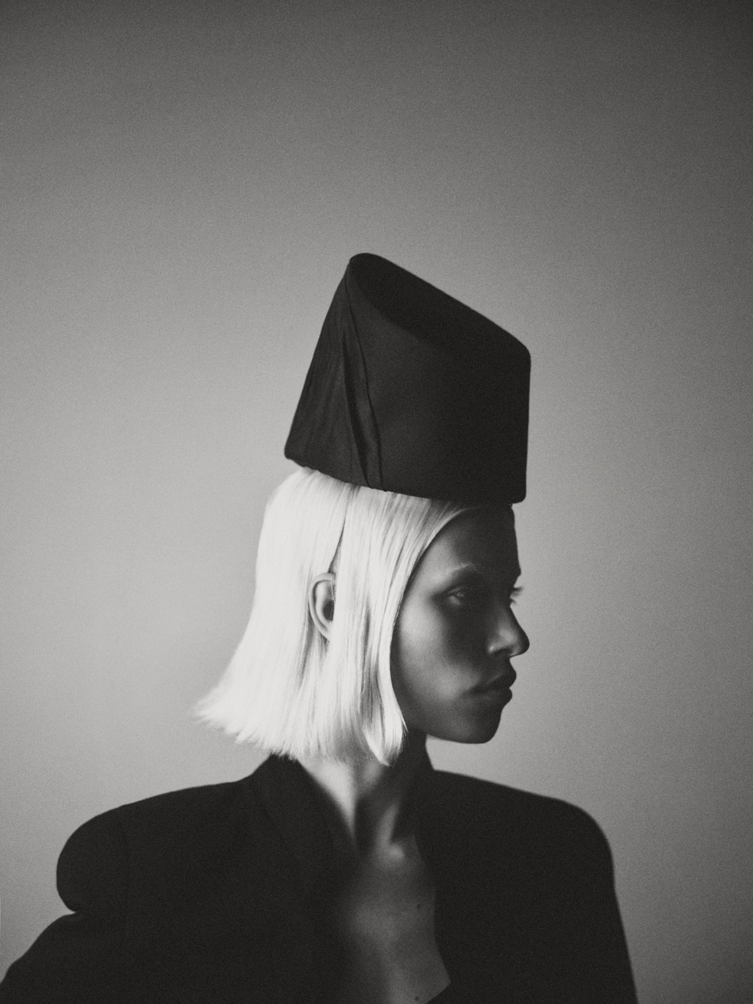Monochrome portrait of a fair-skinned woman in a black jacket and a geometric hat, looking to the side in a studio setting.