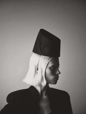 Monochrome portrait of a fair-skinned woman in a black jacket and a geometric hat, looking to the side in a studio setting.