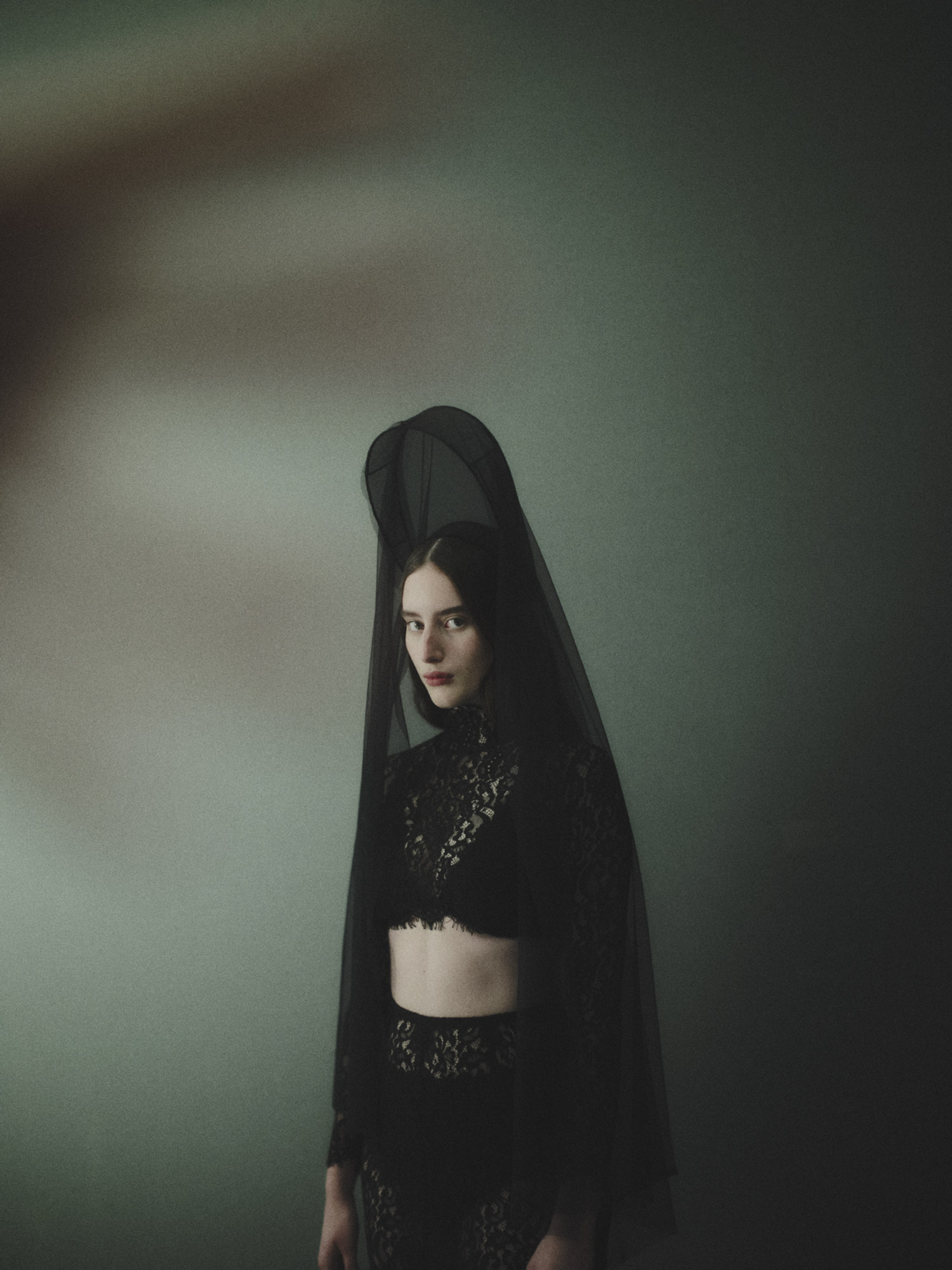 Portrait of a female model in black lace crop top and skirt, wearing a black veil, posed against a teal and grey background.