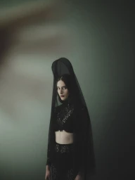 Woman in a black lace crop top and matching veil looking seriously at the camera against a green background