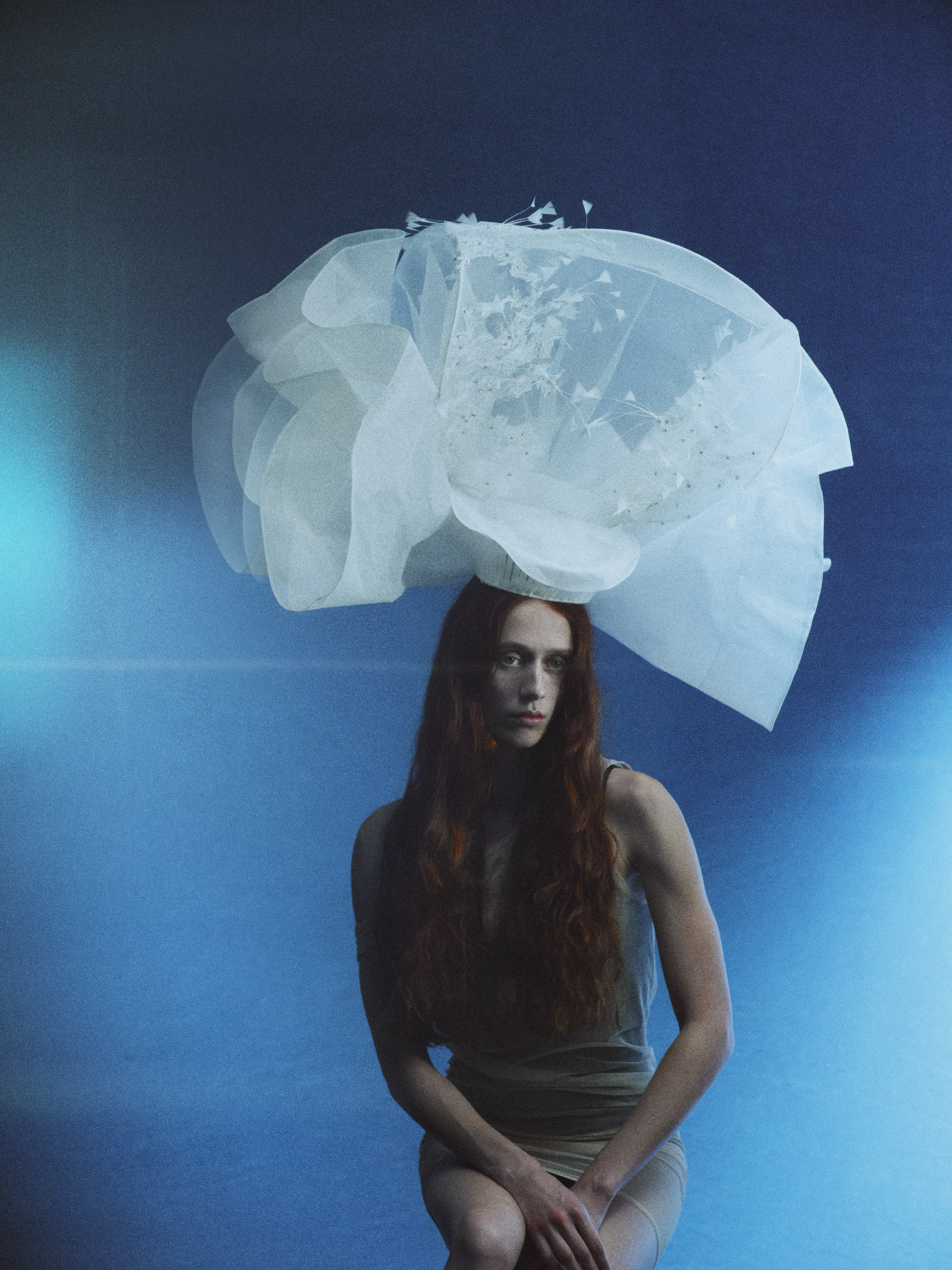 Elegant woman with long red hair poses seated against a blue background wearing an abstract white headpiece in a studio portrait.