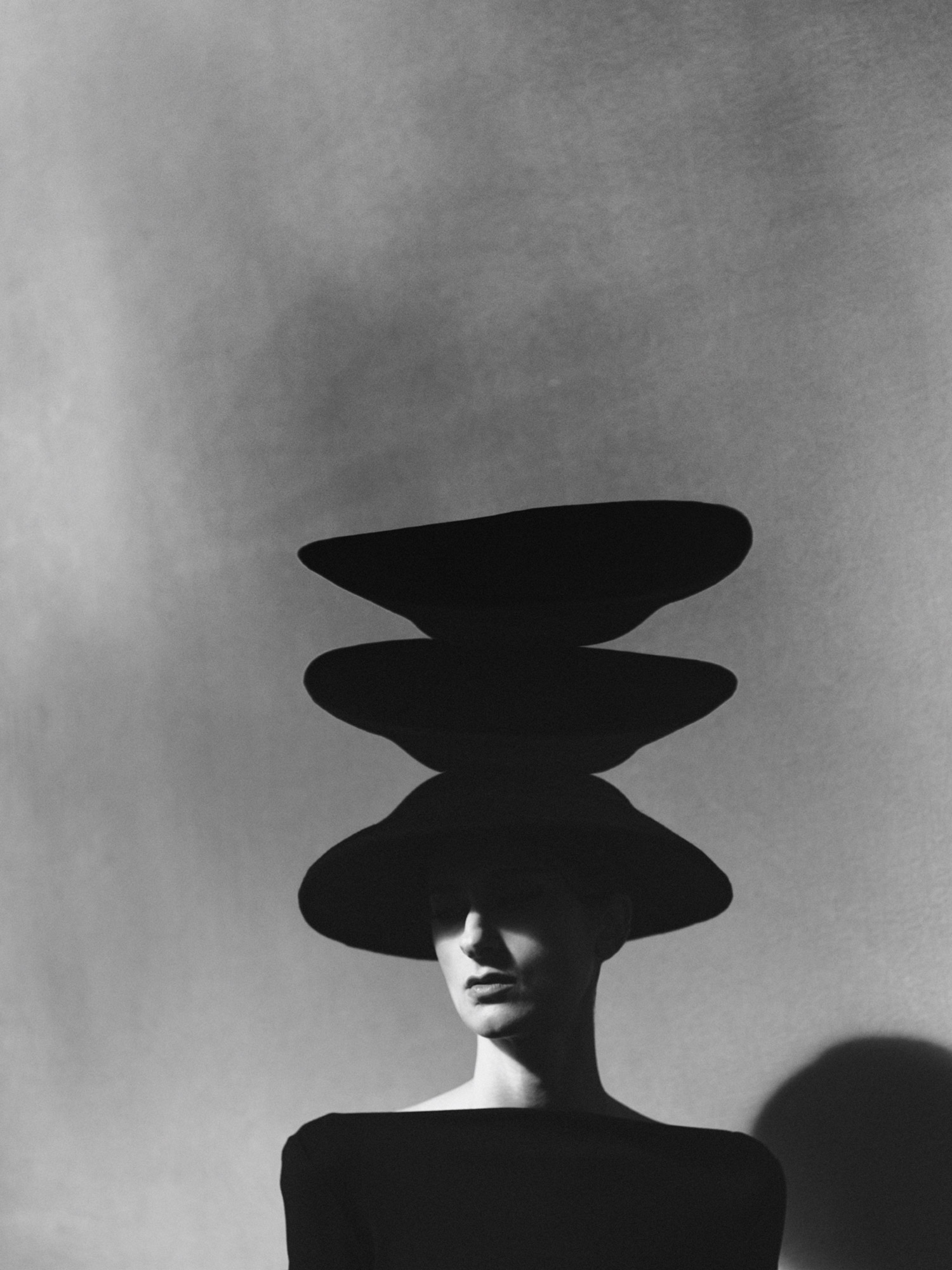 Monochrome portrait of a person wearing three wide-brimmed hats stacked atop their head in a surreal fashion image against a mottled grey backdrop.