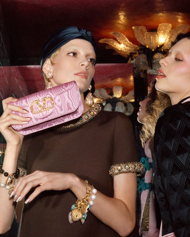 Two fair-skinned adult women with dark head wraps pose indoors with pink clutch purses, wearing jewelry and dark formal dresses.