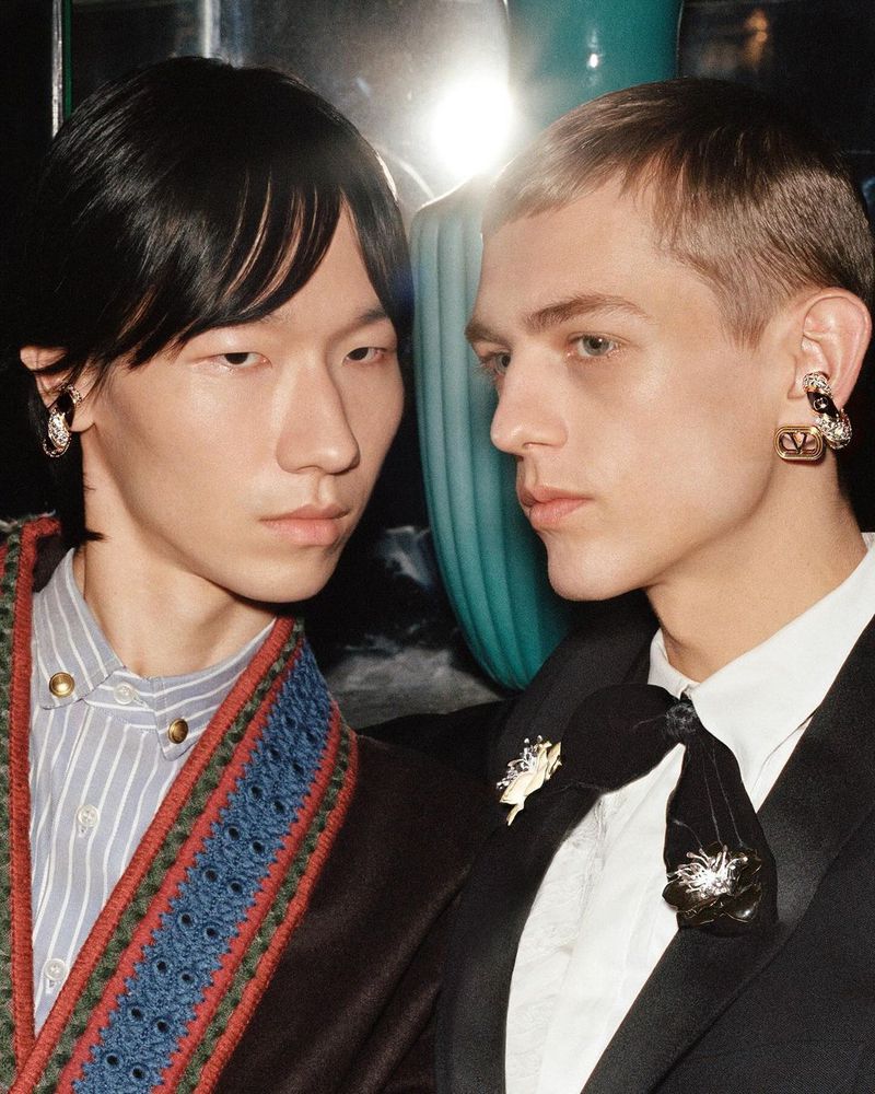 Two men pose indoors with unique earrings and formal attire, showcasing fashion jewelry and classic style in a dimly lit setting.