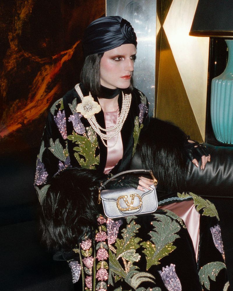 A woman in a turban and ornate coat poses indoors with a designer handbag. The scene features rich textures and tones of blue, gold, and black.