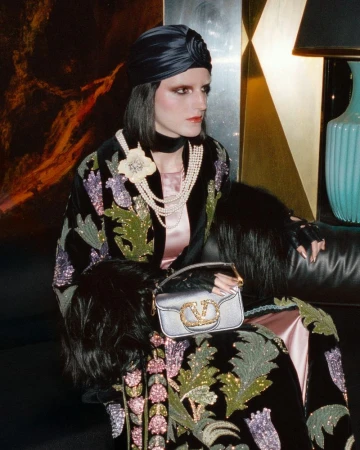 A woman in a turban and ornate coat poses indoors with a designer handbag. The scene features rich textures and tones of blue, gold, and black.