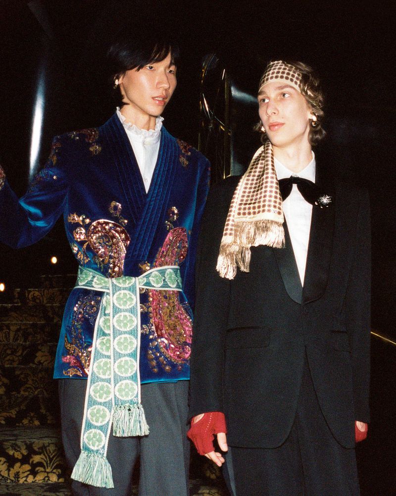 Two stylish men in formal wear pose indoors; one wears a blue velvet jacket with embellishments and a patterned sash, and the other a black tuxedo.