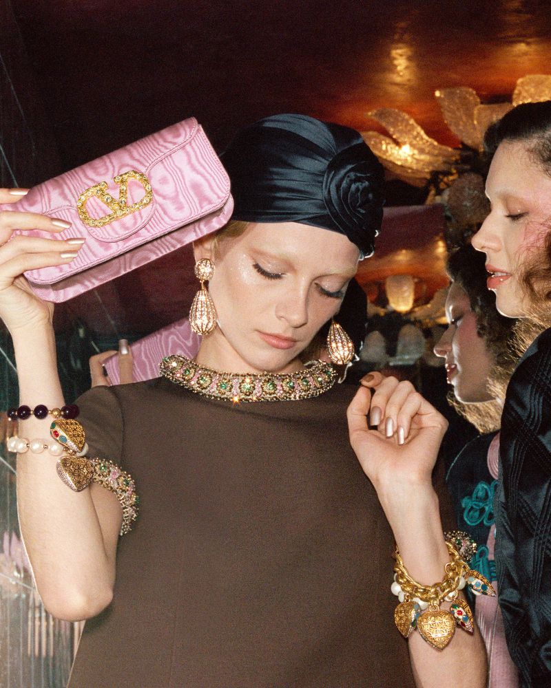 Two fashion models pose with luxury accessories in an indoor setting, showcasing elegant jewelry and a pink Valentino handbag.