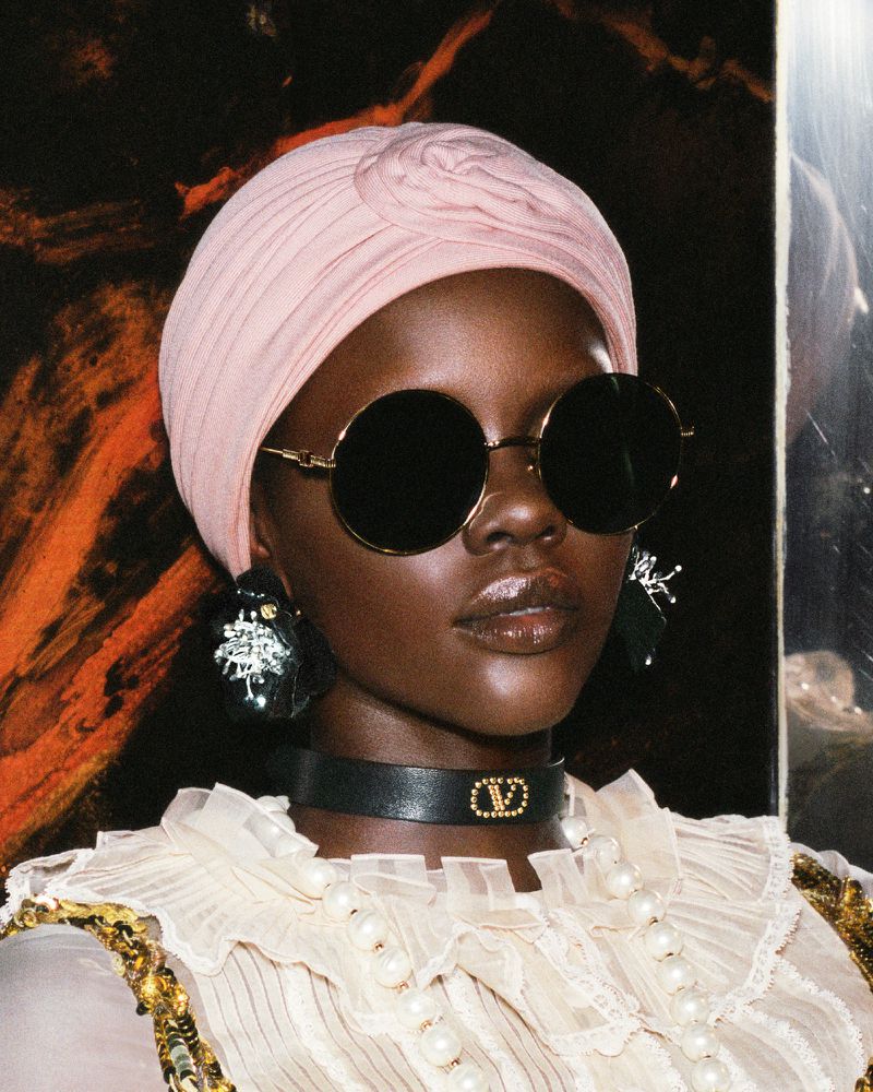 Close-up portrait of a woman with dark skin wearing a pink turban, round sunglasses, black choker necklace and decorative earrings.