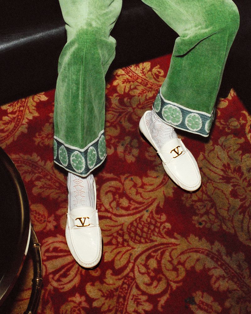 Fashionable composition featuring green velvet pants, white leather loafers with gold emblems, and patterned rug, offering style and luxury.