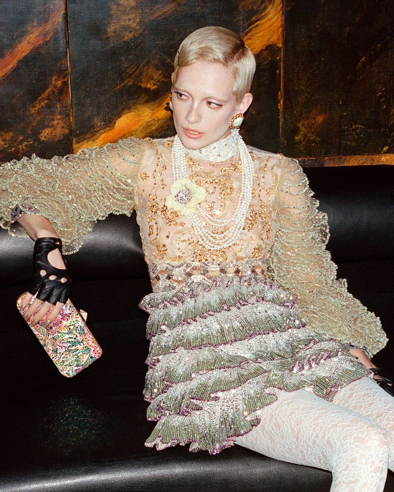 Elegant woman in a sheer, frilly vintage-style dress and pearls sits on a black leather sofa in a dark room, holding a clutch purse.