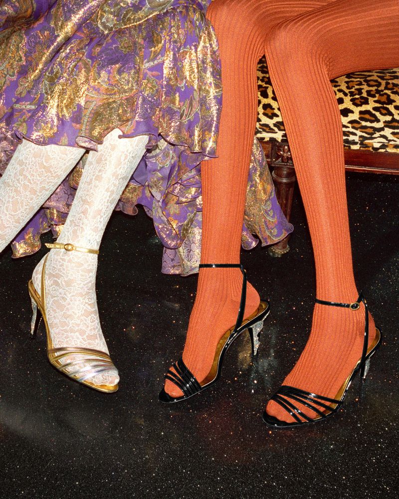 Close-up on two pairs of women's legs wearing stockings, black high heels, and gold accents, against a glossy black background.