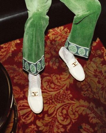 Close-up of green velvet trousers with patterned cuffs, white patterned socks, and white loafers standing on a red ornate patterned rug