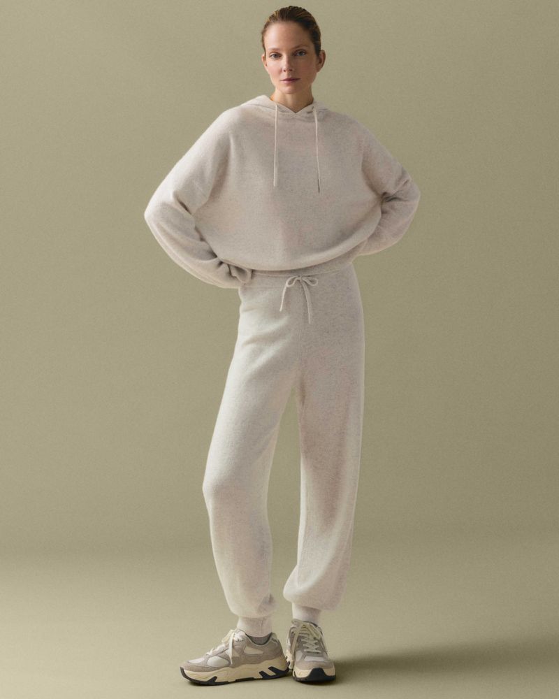 A woman stands in a neutral studio wearing a matching beige hoodie and sweatpants, accessorized with chunky sneakers, in a full shot.