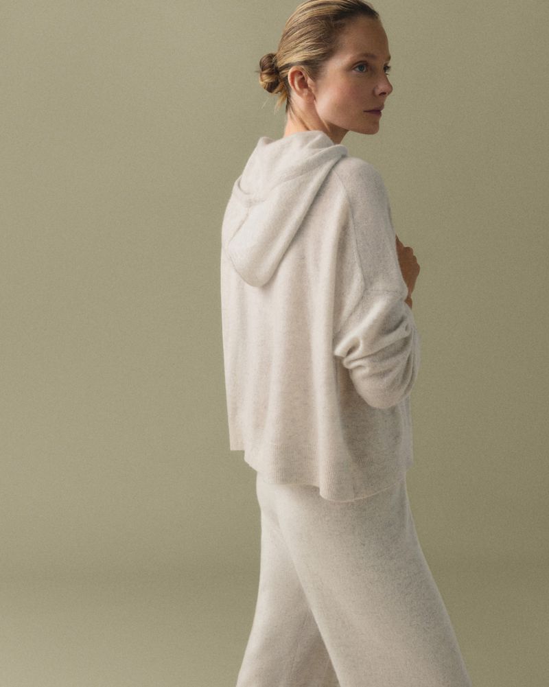 Blond woman in a cream-colored hoodie and pants poses in a studio with neutral tones, looking away from the camera.