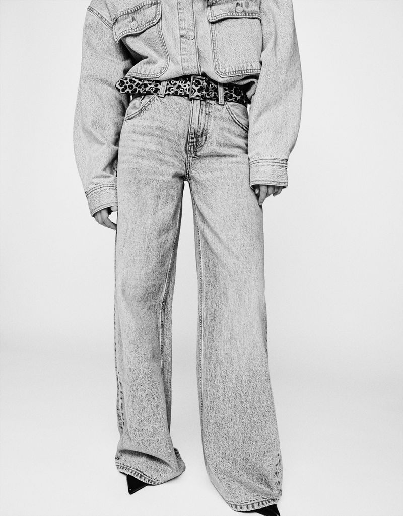 Monochrome fashion shot of a person wearing a denim top and wide-leg jeans with a patterned belt on a white background.