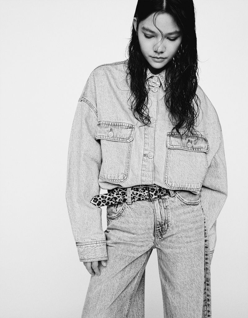 Monochrome portrait of a young adult woman in a light denim jacket and jeans with an animal-print belt, looking down.