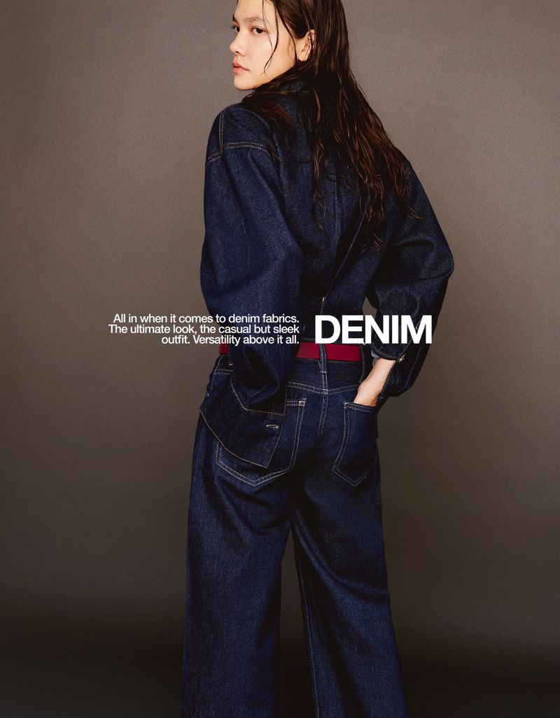 Fashion shot of a woman facing away from the camera wearing a dark blue denim jacket and pants, with wet hair against a grey background.