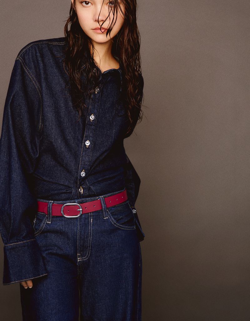 A woman in a denim shirt and jeans is captured in a studio shot against a muted grey background, showcasing a modern fashion look.