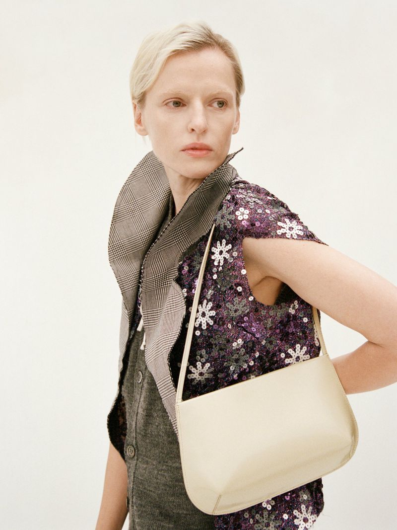 Model wearing a sequined purple top and houndstooth collar holding a cream shoulder bag against a white background