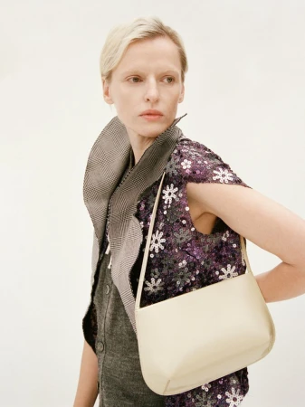 Model wearing a sequined purple top and houndstooth collar holding a cream shoulder bag against a white background