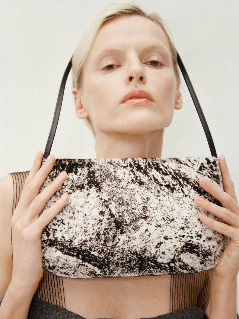 A fair-skinned adult female poses indoors, holding a black and white handbag with black straps against a light background.