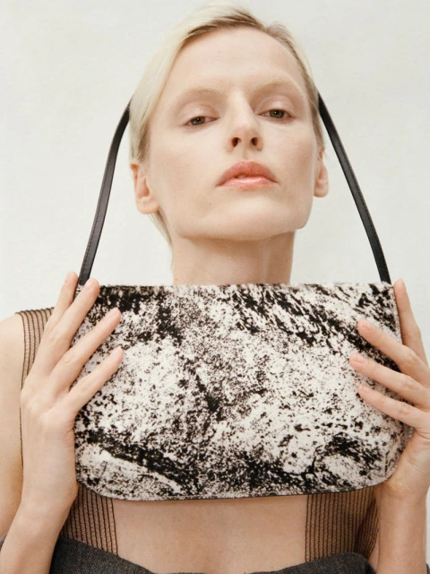 A fair-skinned adult female poses indoors, holding a black and white handbag with black straps against a light background.