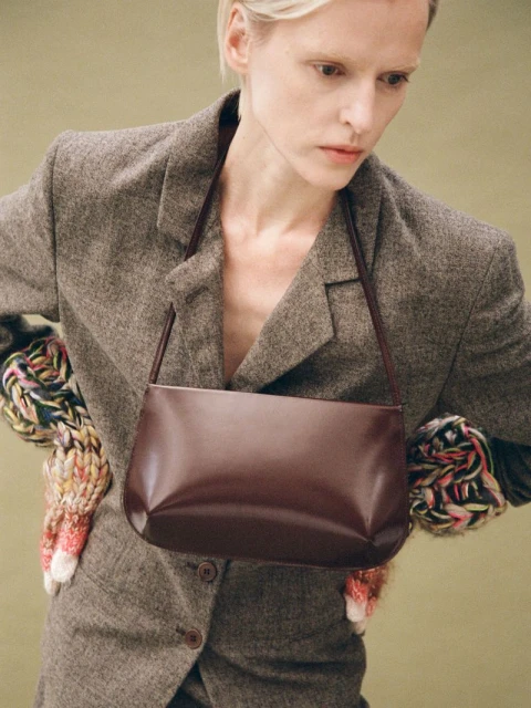 Fashion shot of a slender woman in a grey suit holding a dark brown leather purse with a short shoulder strap in a studio setting.