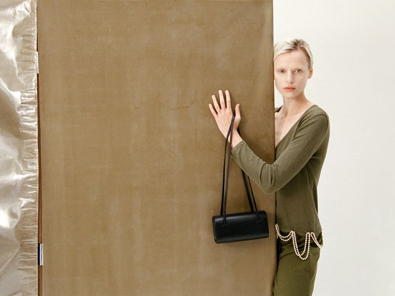 Woman in green outfit with black bag poses against brown panel and metallic backdrop in a fashion-forward neutral studio shot.
