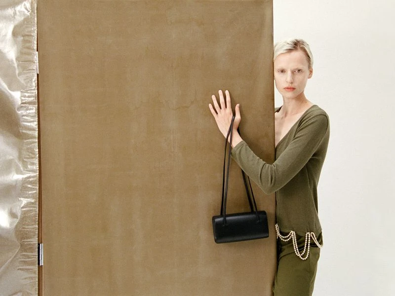 Woman in green outfit with black bag poses against brown panel and metallic backdrop in a fashion-forward neutral studio shot.