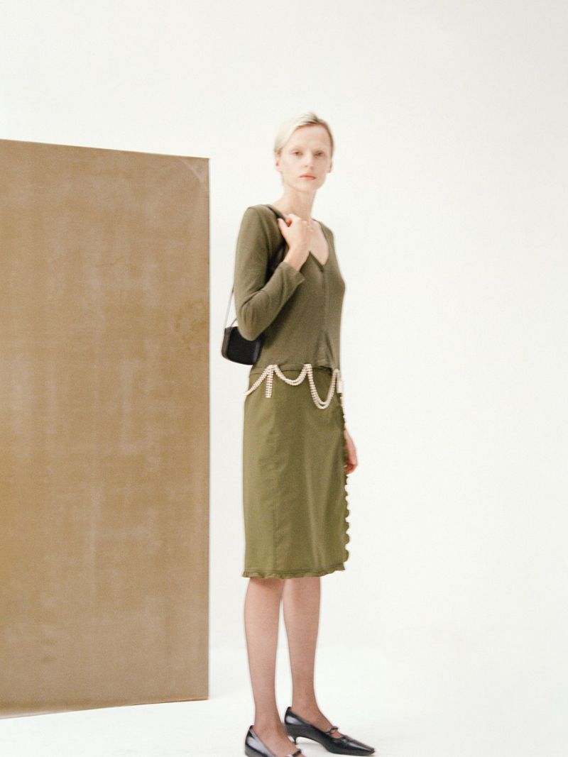 An adult woman in a green dress and dark flats poses in a photo studio with a neutral background and brown accent wall panel.
