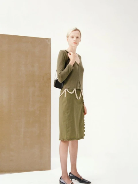 An adult woman in a green dress and dark flats poses in a photo studio with a neutral background and brown accent wall panel.