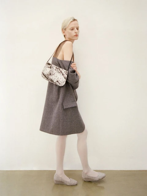 A woman poses in a grey oversized coat with a patterned purse and low heels against a plain light backdrop in a fashion editorial.