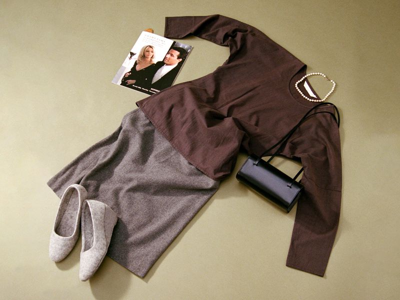 Still life of brown long-sleeved shirt, grey skirt, grey shoes, black purse, and necklace, displayed on a light green surface.