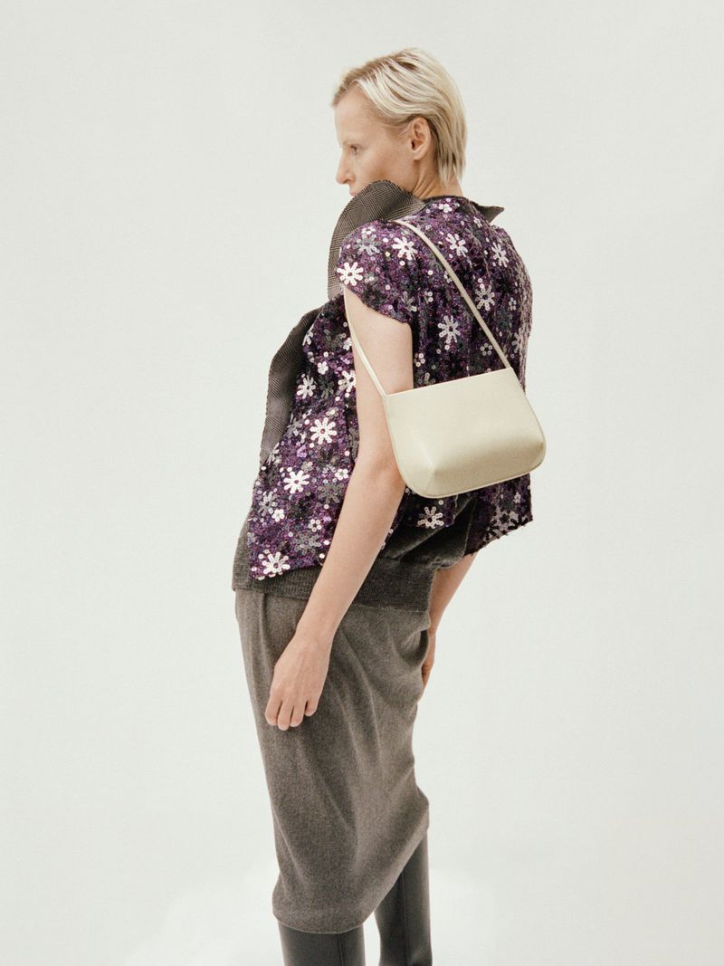 Model wearing a patterned purple floral top and grey skirt carrying a light handbag against a white background