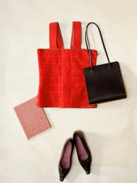 Flat lay of a red tank top, black purse, pink book, and black heels on a cream background, conveying a fashion flat lay composition.
