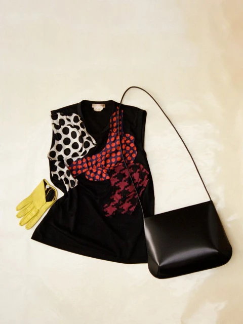 Fashion still life features a black top with graphic patterns, a yellow glove, and a black purse, arranged against a cream backdrop.