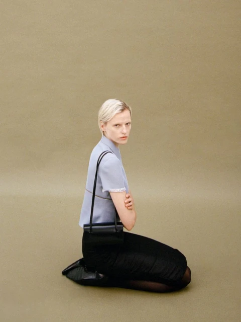 Woman with short blonde hair poses kneeling, wearing a blue top, black skirt, black purse, and black shoes in a studio setting.