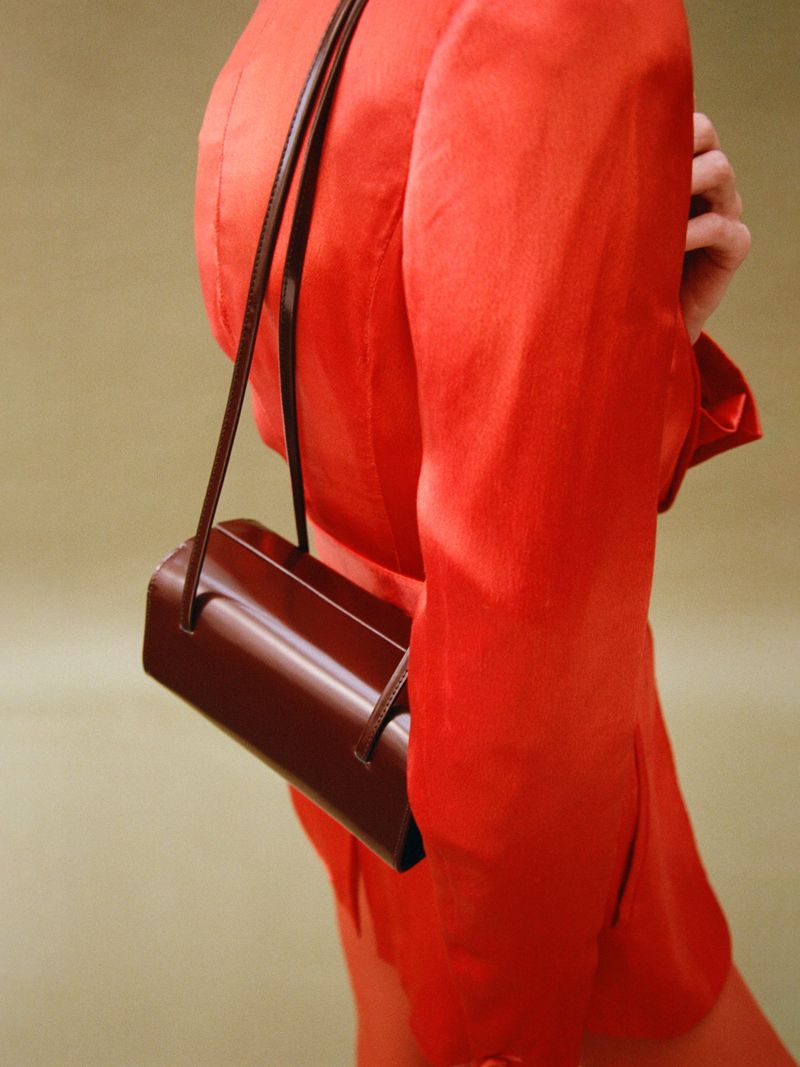 Woman in an orange suit jacket with a brown leather handbag hanging off her shoulder against a soft olive green backdrop.