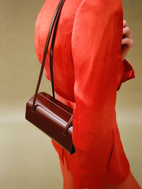 Woman in an orange suit jacket with a brown leather handbag hanging off her shoulder against a soft olive green backdrop.