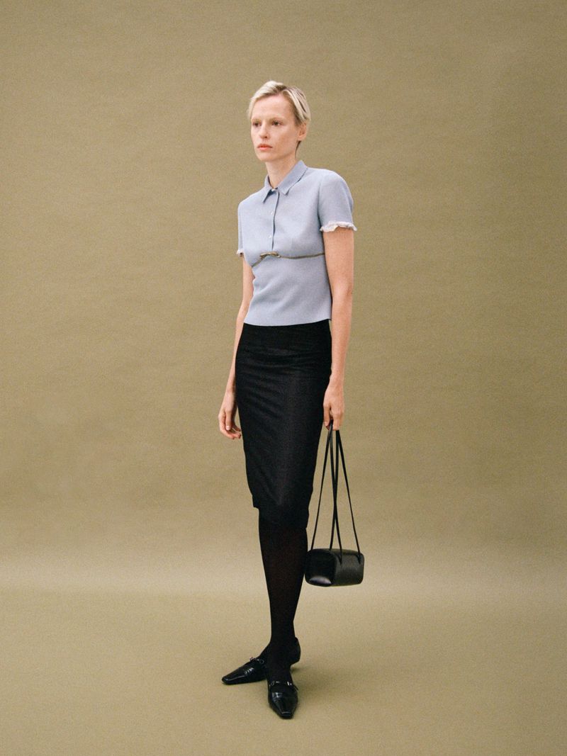 Fashionable adult woman in a collared shirt, dark skirt, black tights and loafers stands in a neutral studio holding a small black purse.