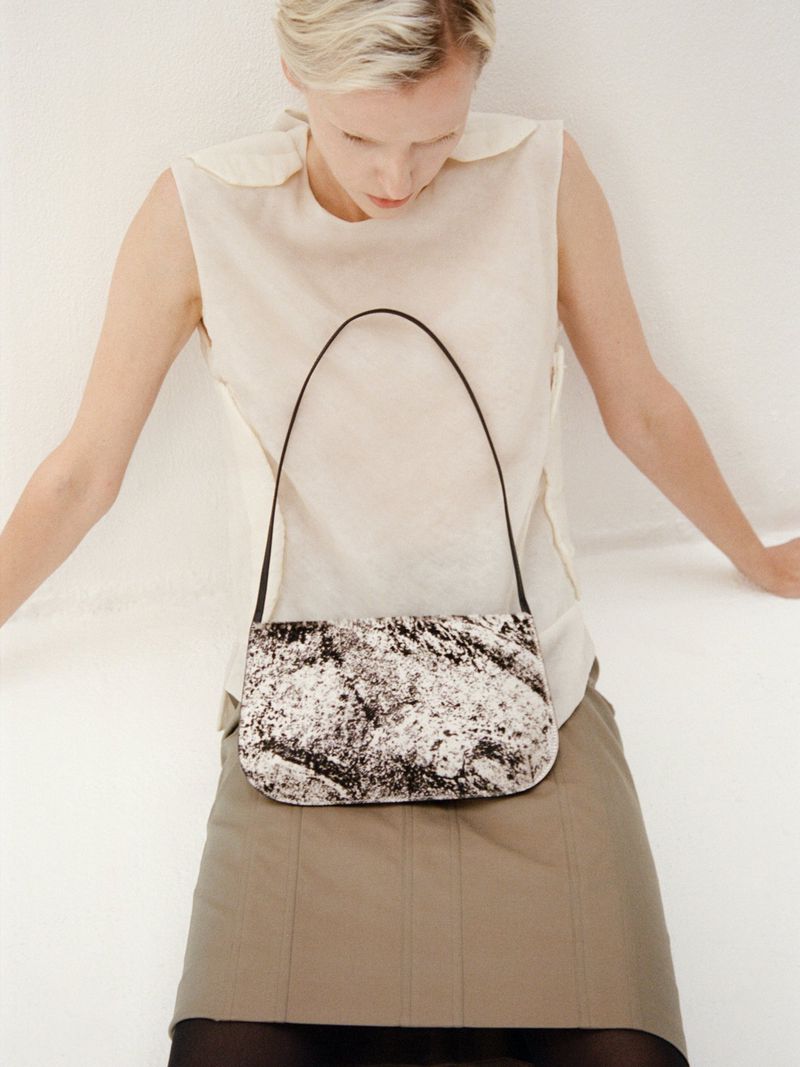 Woman models a black and white patterned handbag in a studio, paired with a cream top and a khaki skirt. Fashion editorial.