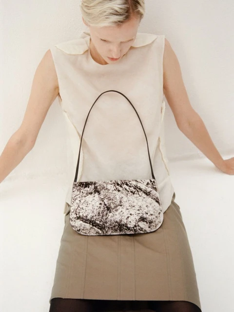 Woman models a black and white patterned handbag in a studio, paired with a cream top and a khaki skirt. Fashion editorial.