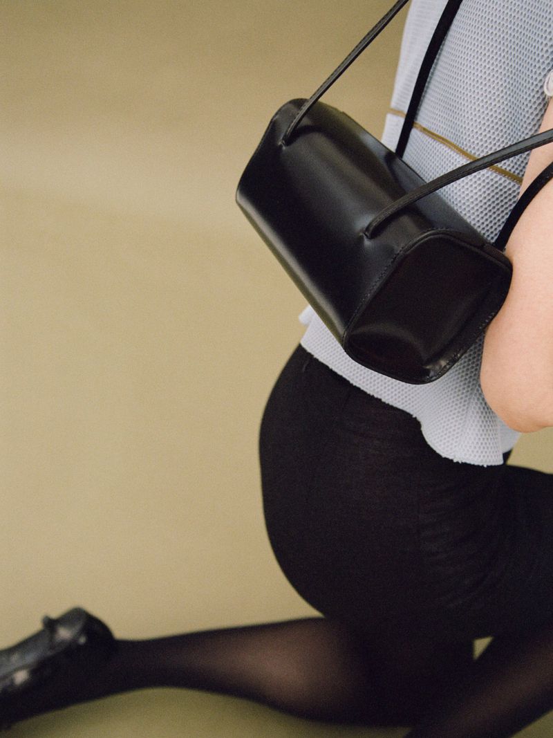 A black leather purse with a cylindrical shape and thin straps is held by an adult female model kneeling in black tights.