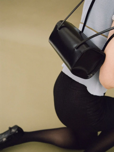 A black leather purse with a cylindrical shape and thin straps is held by an adult female model kneeling in black tights.