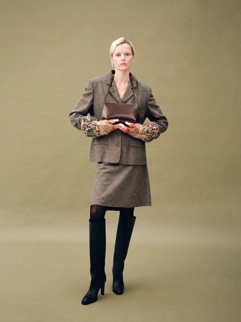 Model in a brown tweed suit holding a dark leather handbag against a solid olive green studio backdrop.