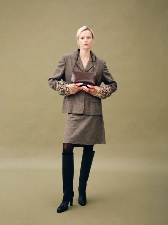 Model in a brown tweed suit holding a dark leather handbag against a solid olive green studio backdrop.